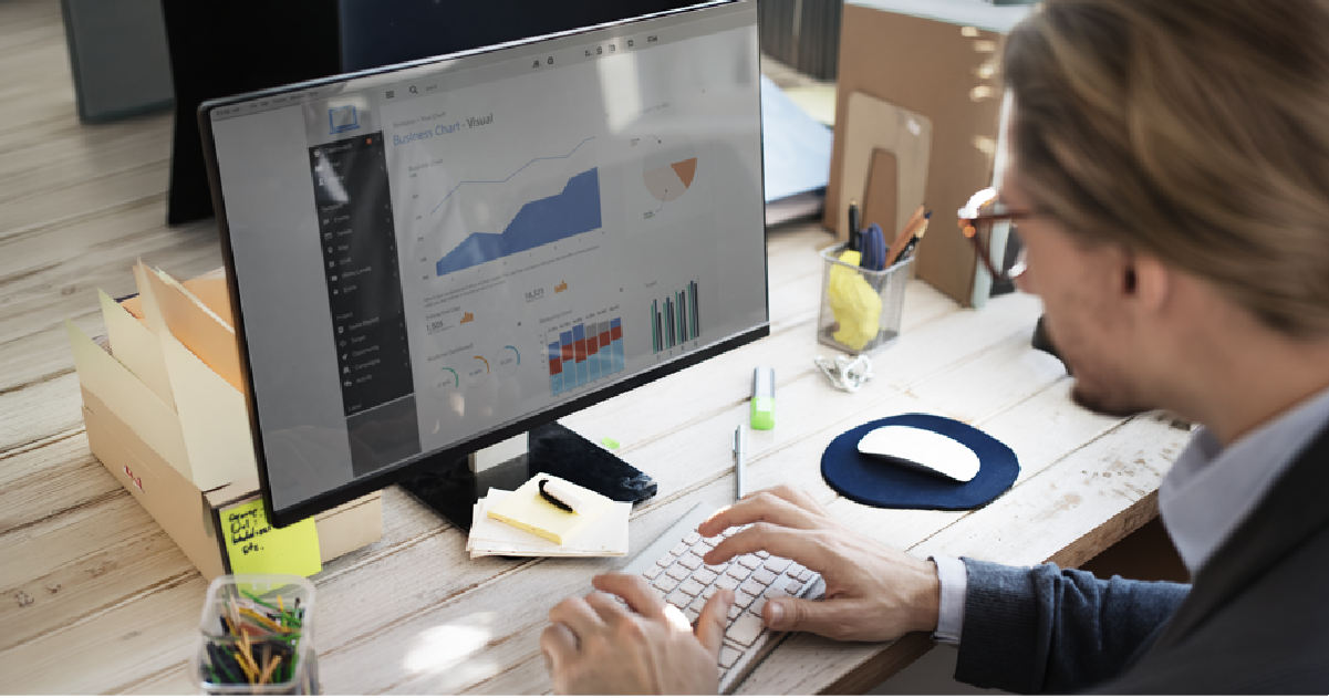 Person typing at a desk with a monitor displaying business charts and graphs