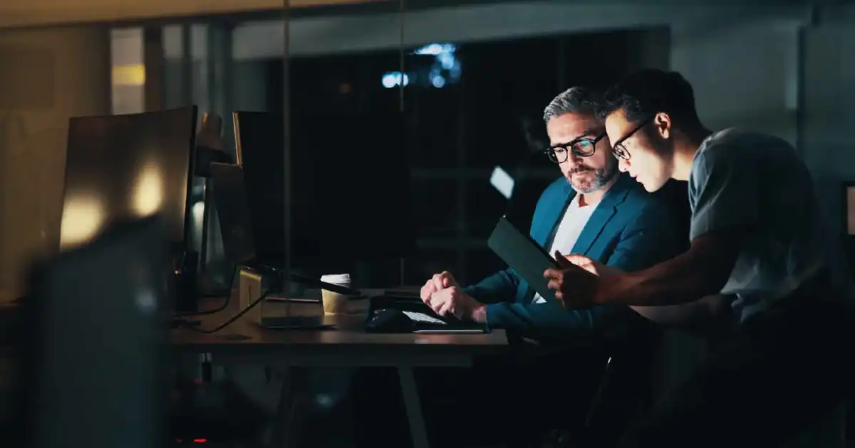 Two men collaborate at a dimly lit desk, examining a tablet beside computer monitors at night.