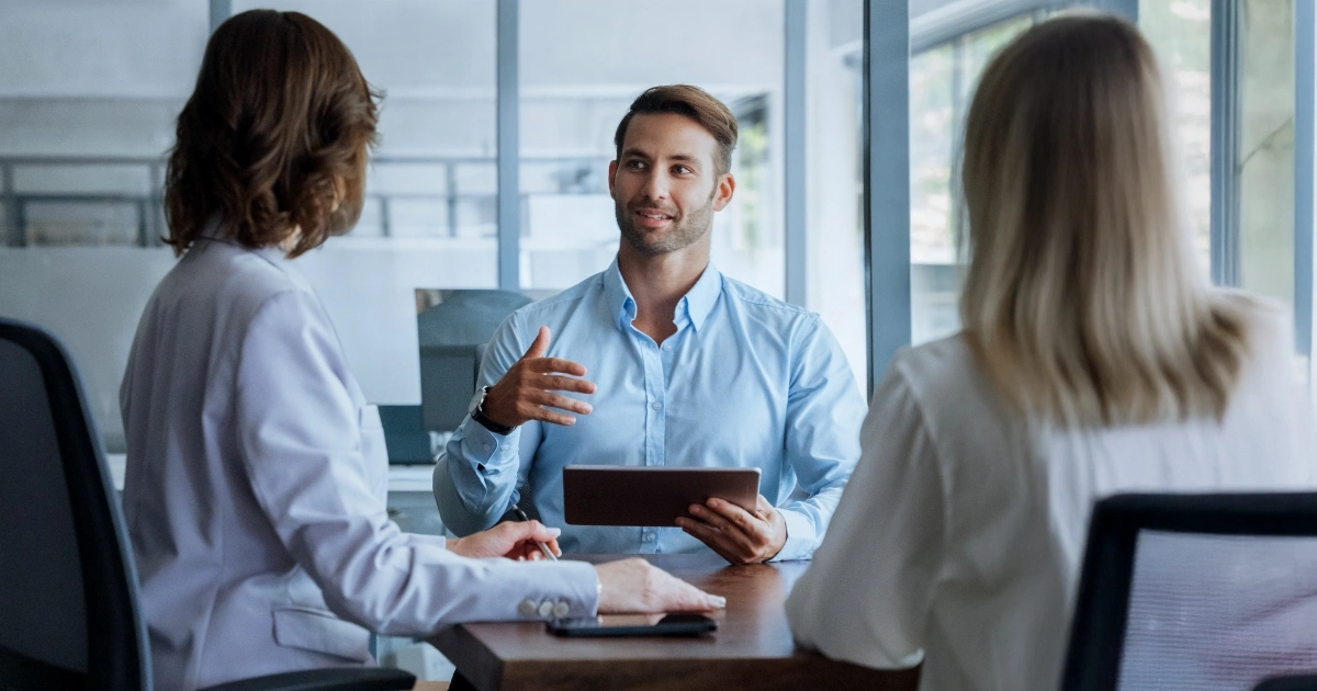 Three colleagues in a meeting: a man in a light blue shirt gestures while holding a tablet, facing two women in an office conference room.