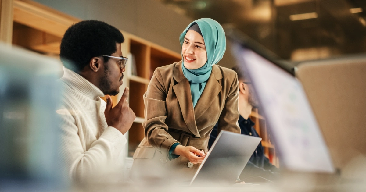 Woman in teal hijab and beige blazer helping a man with a laptop in a modern coworking space