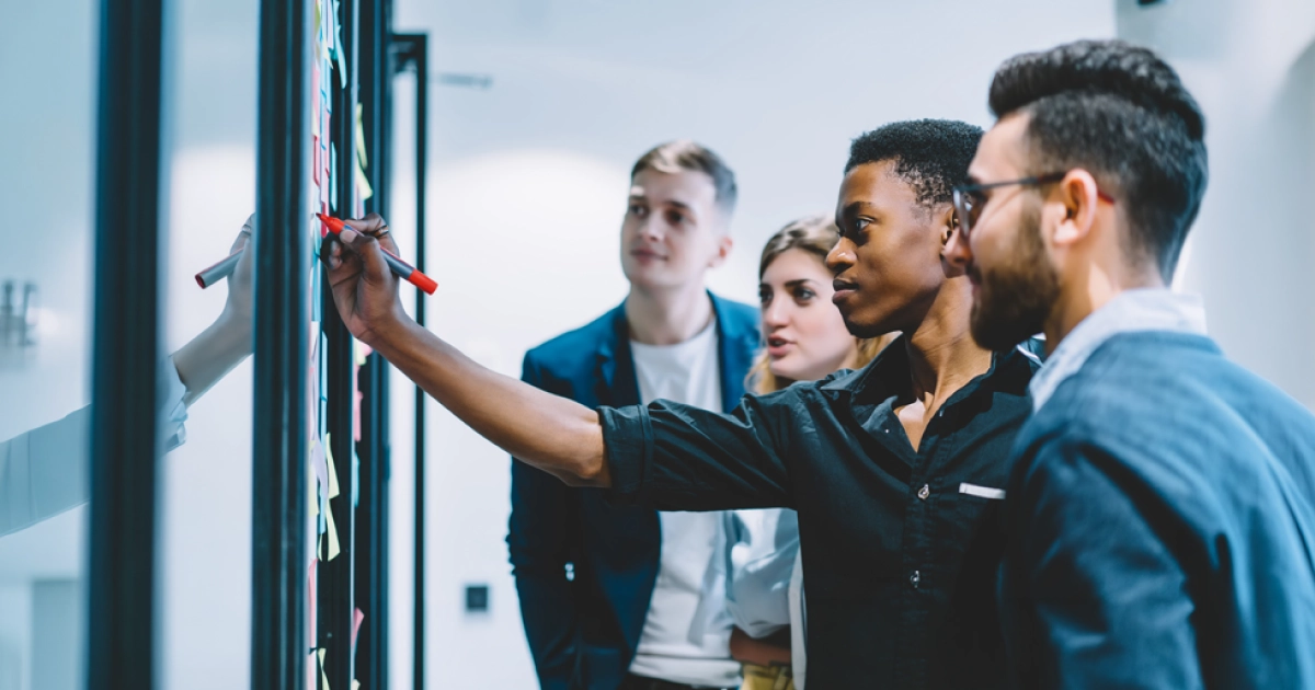 Diverse group of professionals stands by a glass wall covered with sticky notes as one person writes with a red marker on the board.
