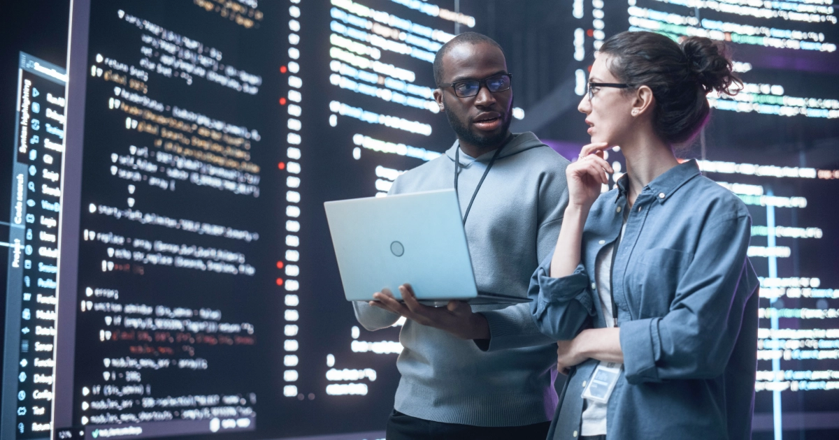 Two programmers collaborate, reviewing code on a laptop in a high-tech data-center or studio with large code displays behind them.