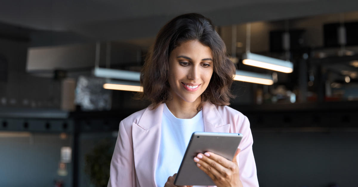 Smiling woman in a light pink blazer uses a tablet in a modern office setting.