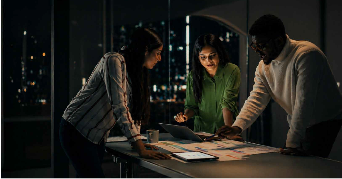 Three coworkers review documents on a table with a laptop and tablet in a nighttime office.