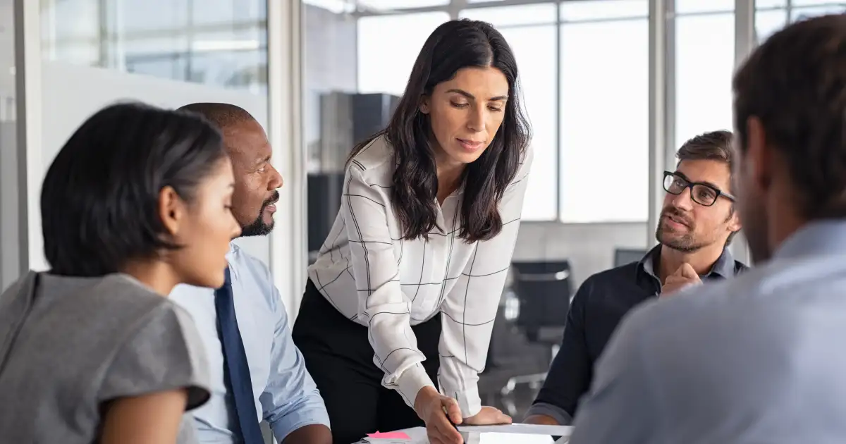 Woman leads a meeting, leaning over a table to discuss documents with four colleagues in a bright office setting.