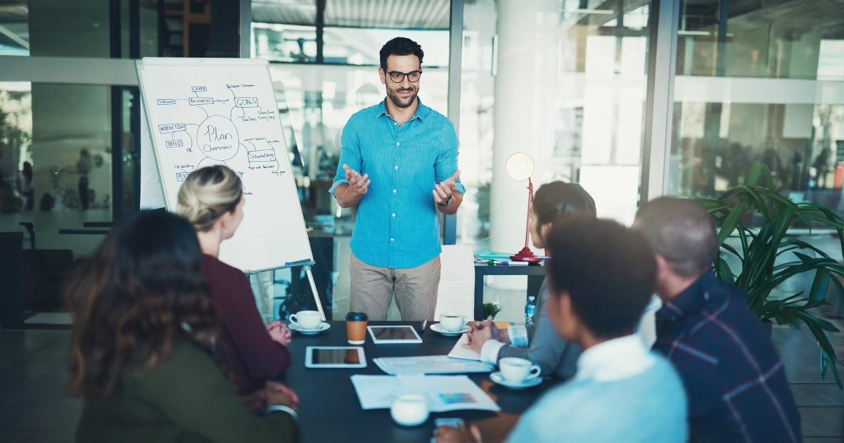 Presenter in a blue shirt leads a planning workshop as colleagues listen around a conference table with tablets and cups nearby near a whiteboard with diagrams.