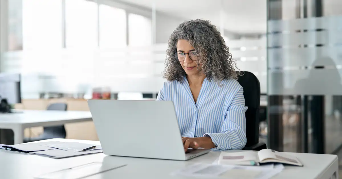 Smiling woman with curly gray hair wearing a blue striped shirt works on a laptop at a bright office desk.**