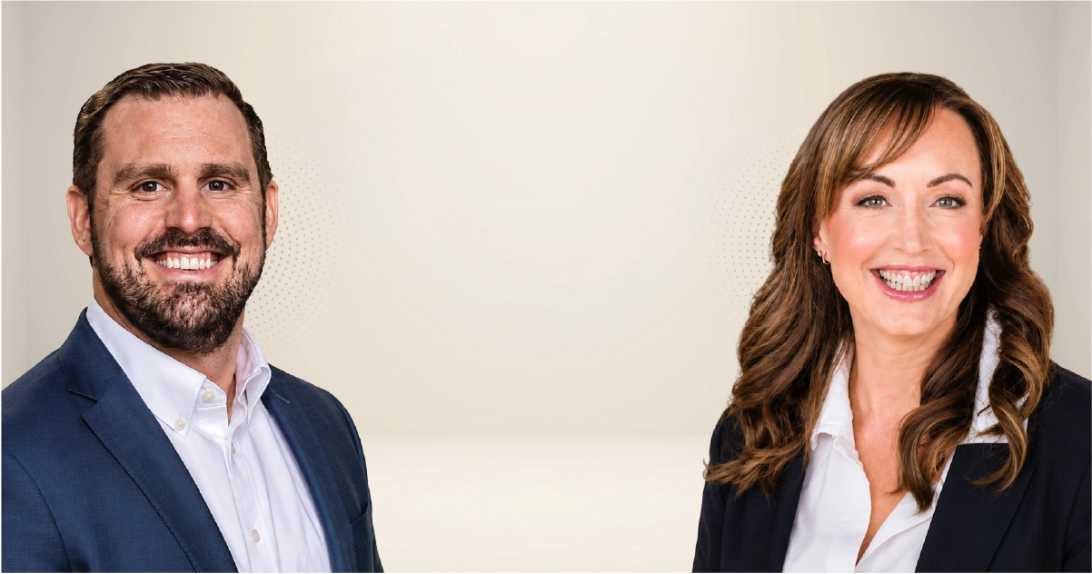 Two business professionals, a man and a woman, smile at the camera in a neutral studio setting.