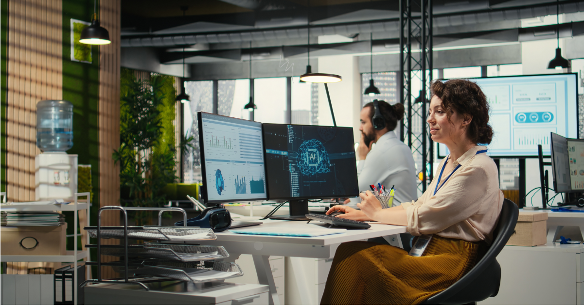 Woman at a white desk with three monitors displaying charts and a neural/brain graphic in a bright open-office setting.