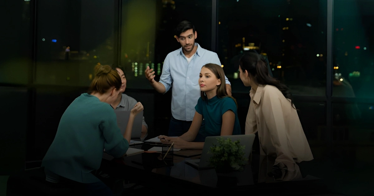 Man standing and explaining something to four colleagues around a conference table at night, laptops open on the table.