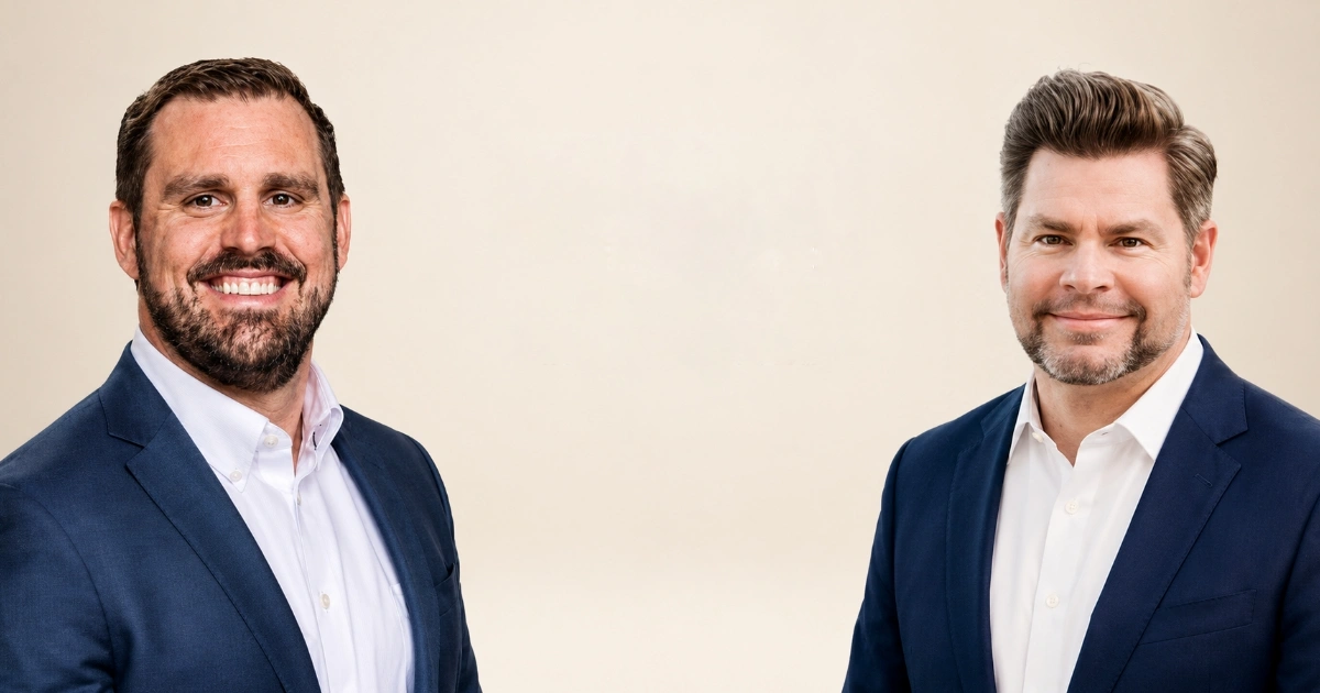 Two men in navy suits and white shirts smile at the camera against a light beige background on a professional portrait.