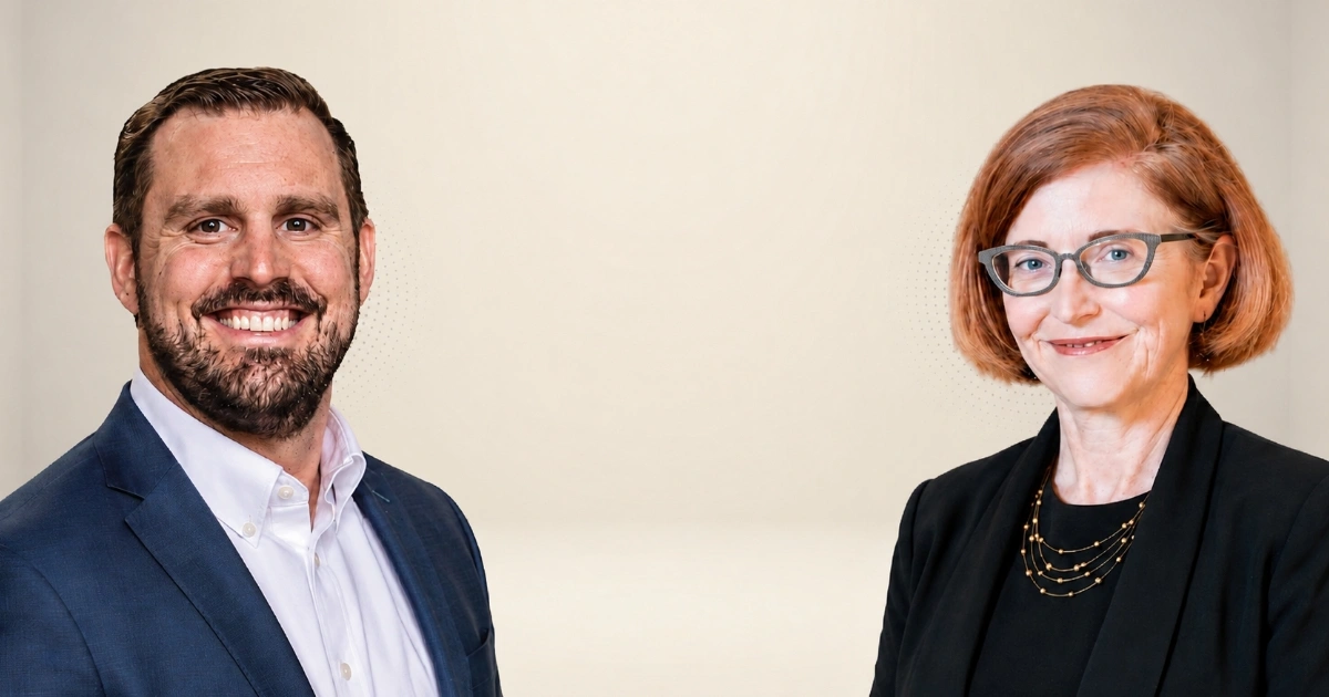 Two professionally dressed individuals smiling at the camera: a man in a navy suit and a woman with red hair and glasses.