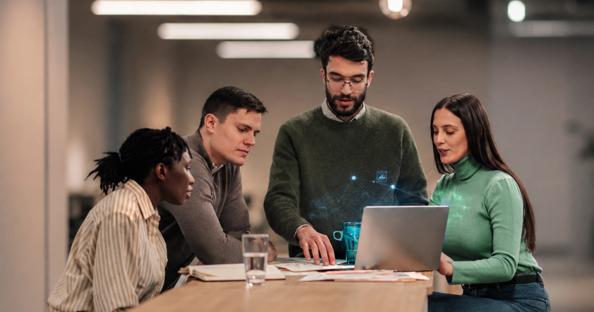 Four colleagues collaborate around a desk, looking at a laptop with a blue digital network projection.