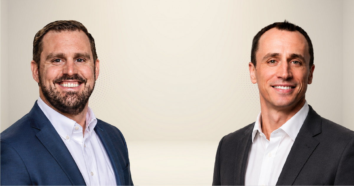 Two men in business suits smiling at the camera against a light neutral background.
