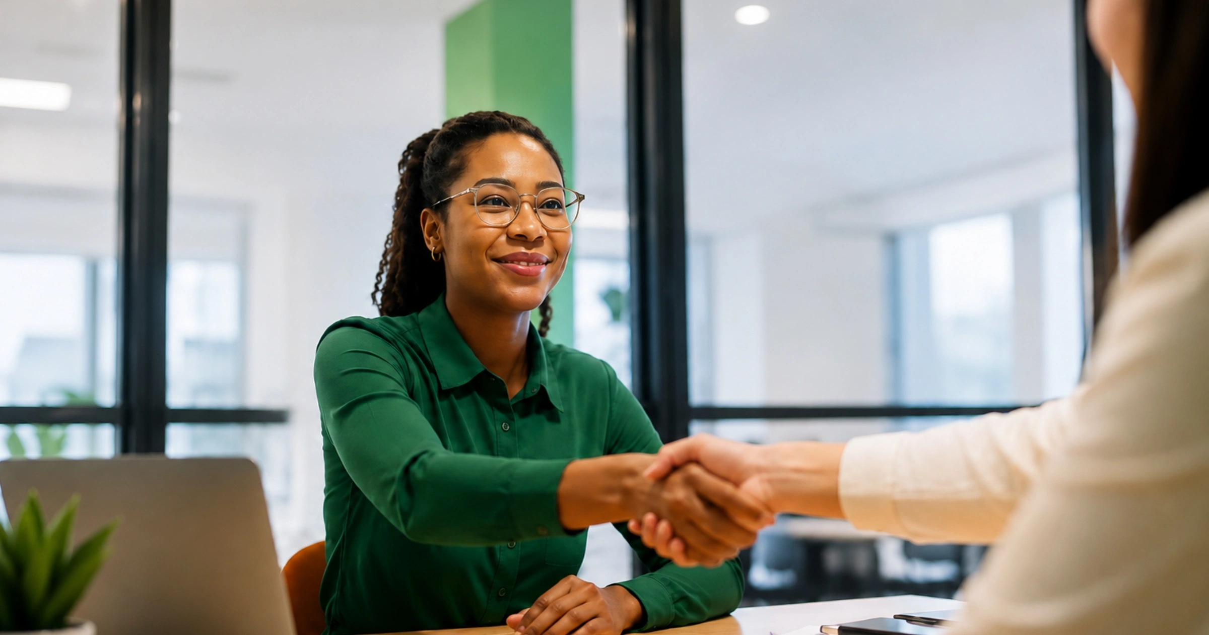 Two colleagues shake hands across a desk in a bright office, smiling after a meeting