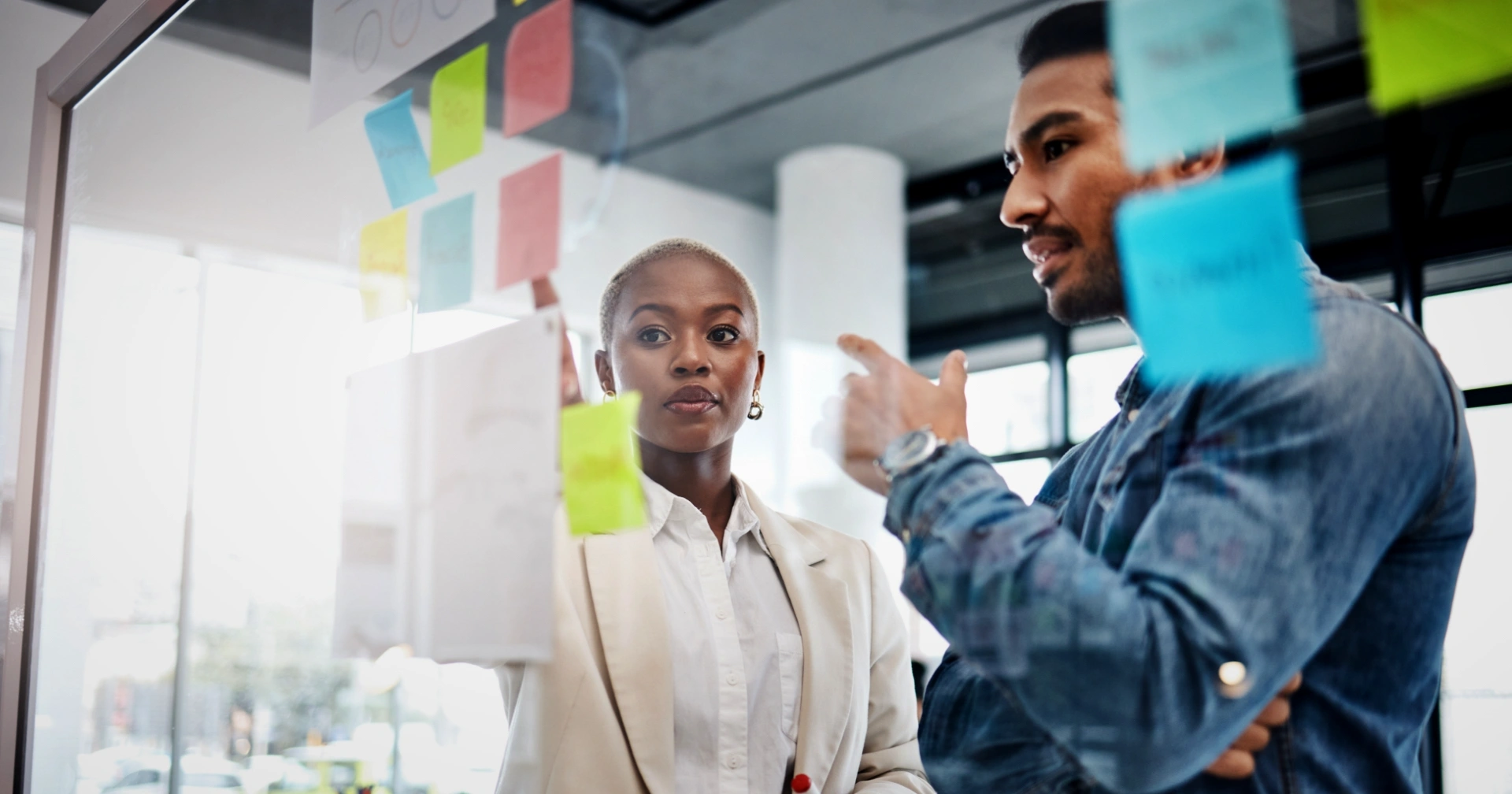 Two coworkers discuss colorful sticky notes on a glass wall during a brainstorming session in a bright office.